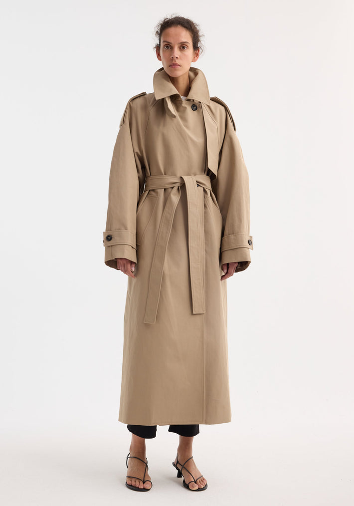 Model in Oversized classic trench | clay, featuring double-breasted design, epaulettes, belted waist, and side pockets, over black cropped trousers and strappy sandals.