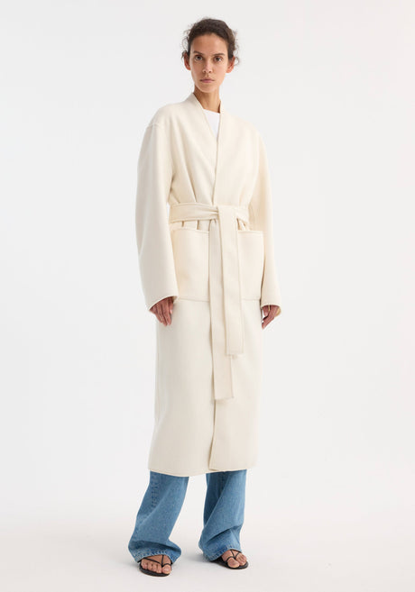 Woman in off-white double-faced kimono style coat with patch pockets, tied at waist. Paired with light-blue jeans and black sandals. 100% wool composition.