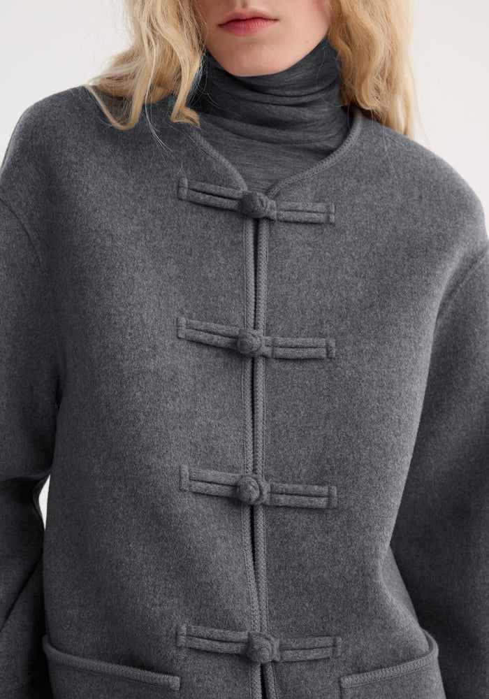 Mandarin double-faced wool jacket with pankou closure, embroidered edges, and patch pockets. Focus on texture and knotted fastenings, worn over a matching turtleneck.