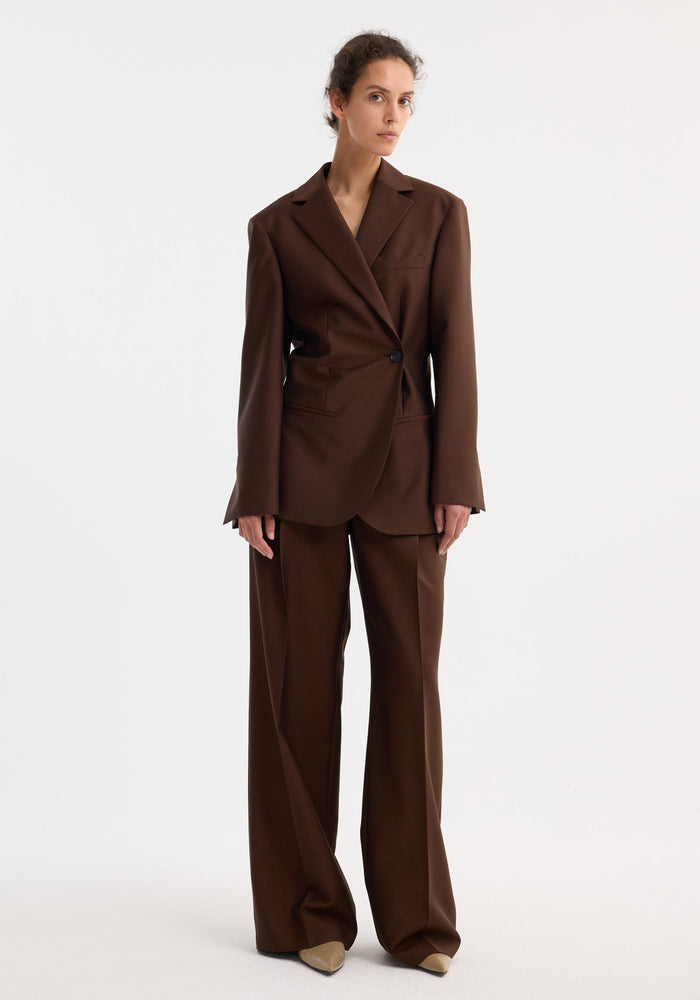 Model wears Dual tone piqué overlap suit jacket in mahogany, featuring a tailored fit and dual-tone fabric, paired with matching wide-leg trousers and pointed-toe shoes.