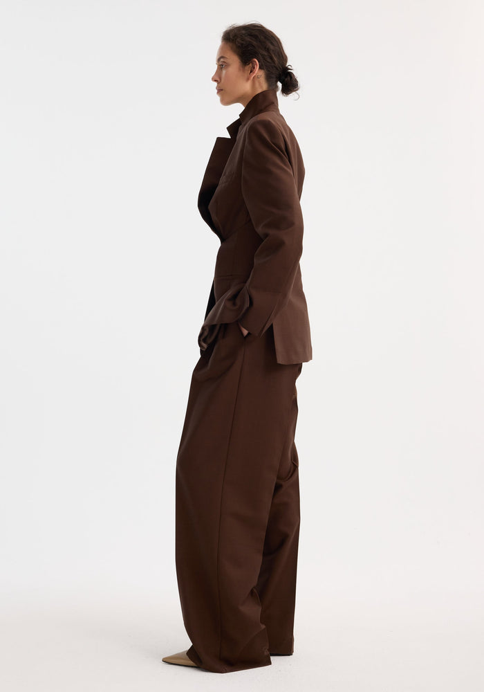 Woman in profile wearing a Dual tone piqué overlap suit jacket in mahogany with matching wide-leg trousers, hands in pockets, conveying a relaxed, contemporary style.