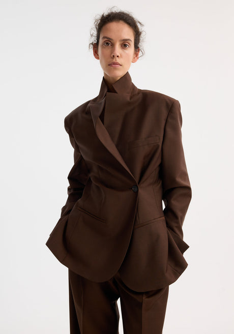 Model wears Dual tone piqué overlap suit jacket in mahogany, featuring a tailored fit and dual-tone fabric, paired with matching wide-leg trousers and pointed-toe shoes.