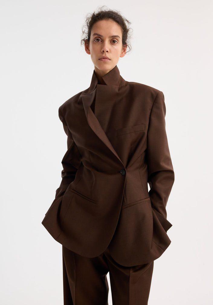 Model wears Dual tone piqué overlap suit jacket in mahogany, featuring a tailored fit and dual-tone fabric, paired with matching wide-leg trousers and pointed-toe shoes.