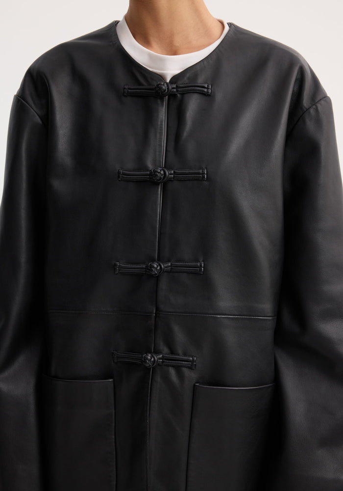 Mandarin leather jacket in black with pankou closures, collarless design, and patch pockets. Worn over a white crew-neck shirt, showcasing its sleek style.