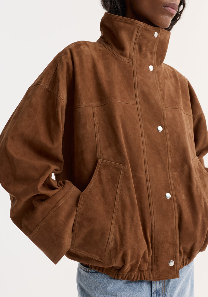 Reinvented suede jacket in cognac with a stand-up collar, drop shoulders, patch pockets, and snap-button closures, modeled with hands in pockets.
