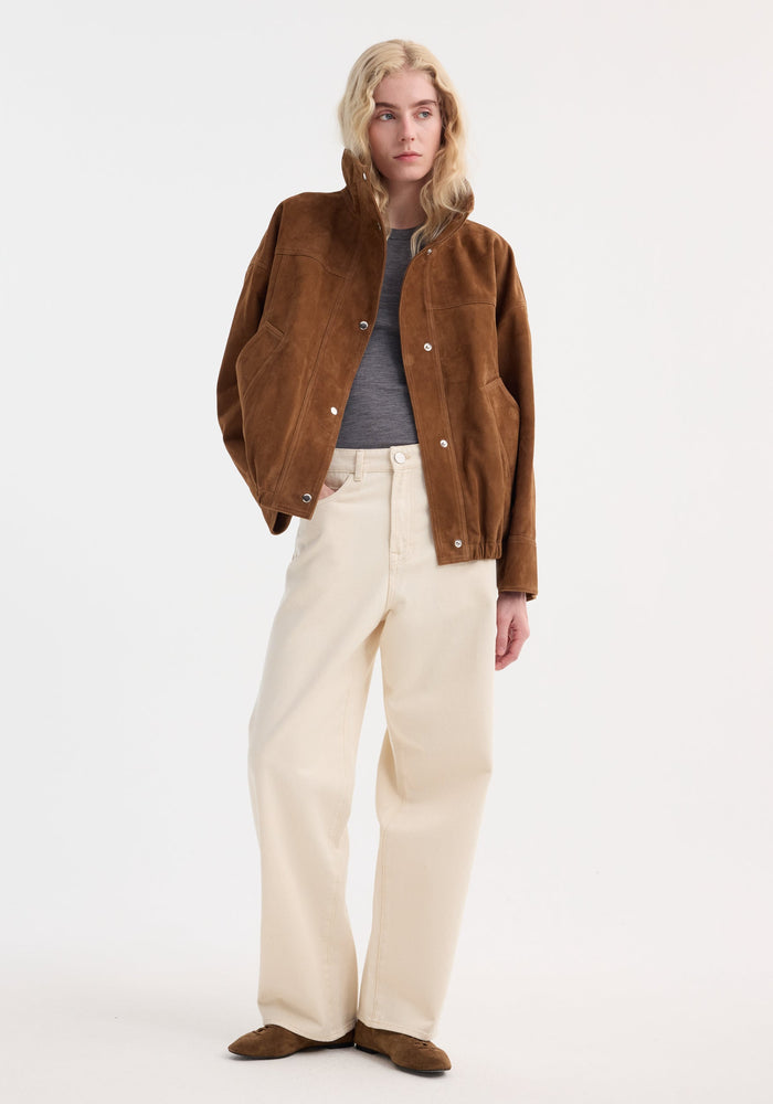 Young adult in a Reinvented suede jacket | cognac, with relaxed bomber style, stand collar, and snap buttons, paired with wide-leg jeans and suede flats.