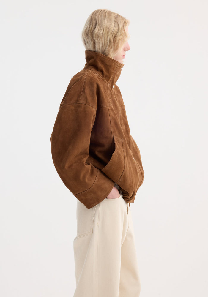 Reinvented suede jacket in cognac, featuring a high stand-up collar and relaxed fit, worn with wide-leg trousers. Model hands in pockets, side profile view.