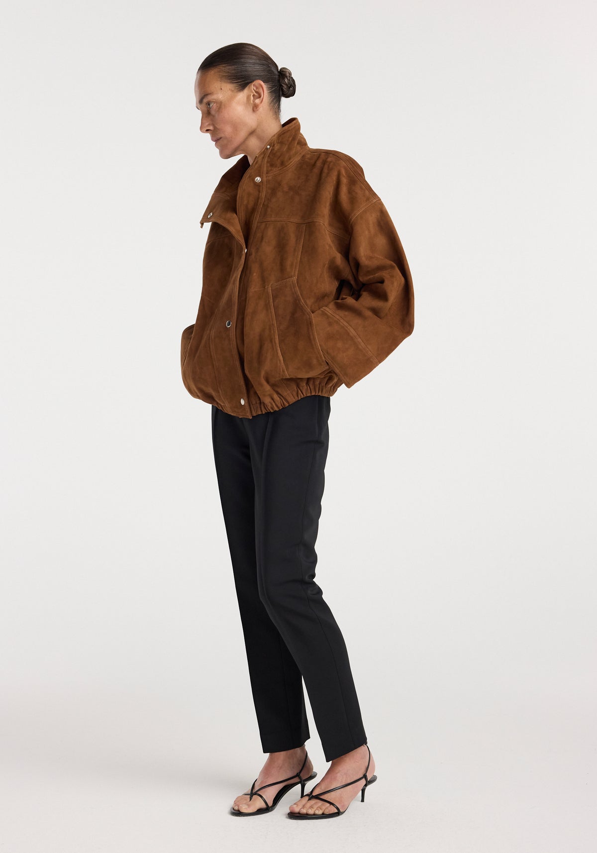 Woman wearing a Reinvented suede jacket | cognac in a loose fit with a high collar, paired with black fitted pants and strappy black heels against a white background.