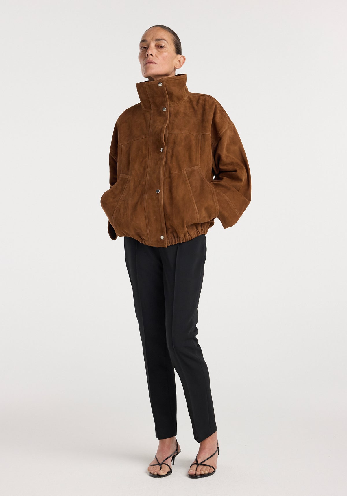 Woman wearing a high-collar, loose-fit Reinvented suede jacket | cognac paired with slim black pants and open-toe heels against a plain light background.