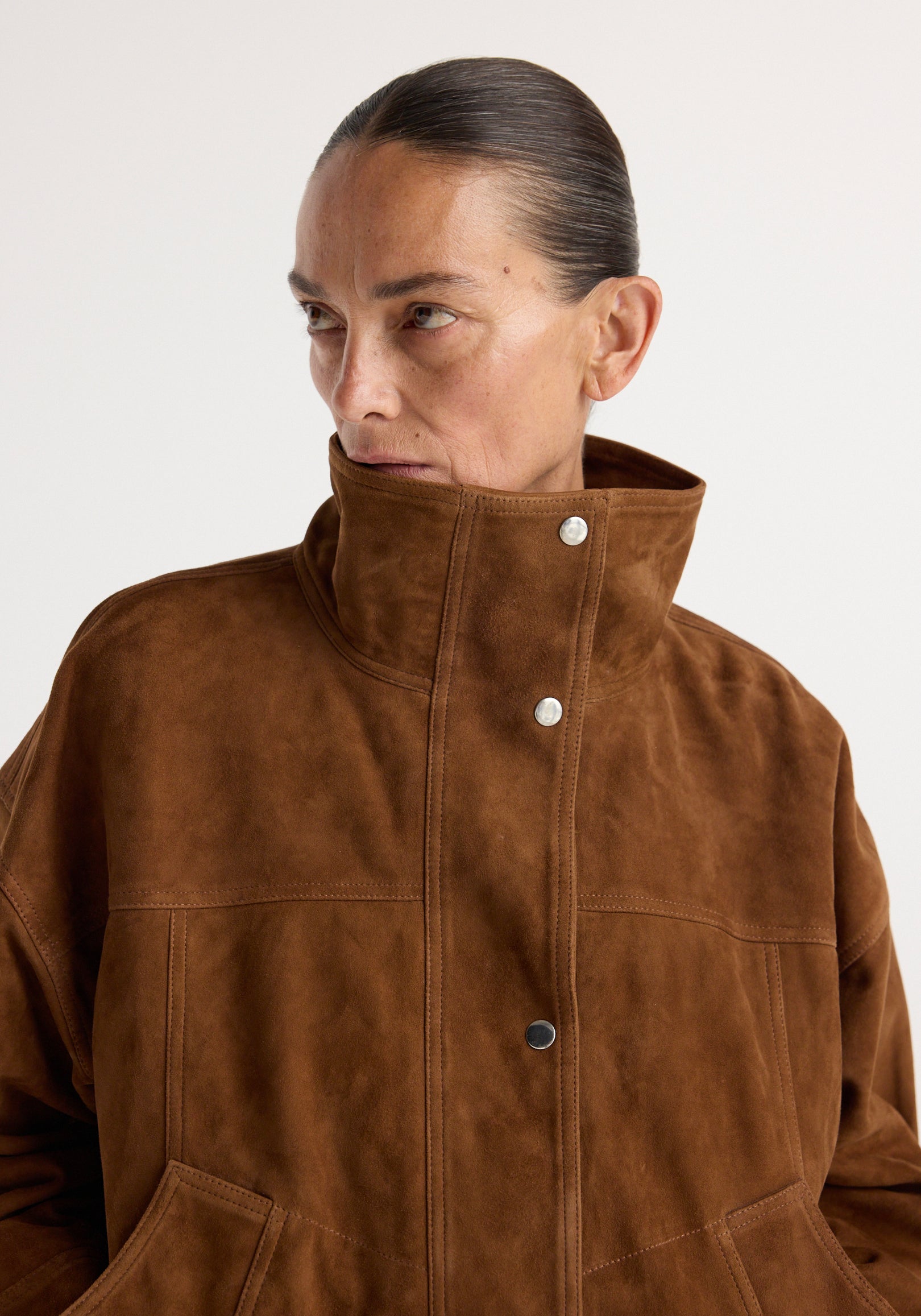 Close-up of a woman wearing a high-collared Reinvented suede jacket | cognac, featuring a rich brown suede material with snap button closures and visible stitching details.