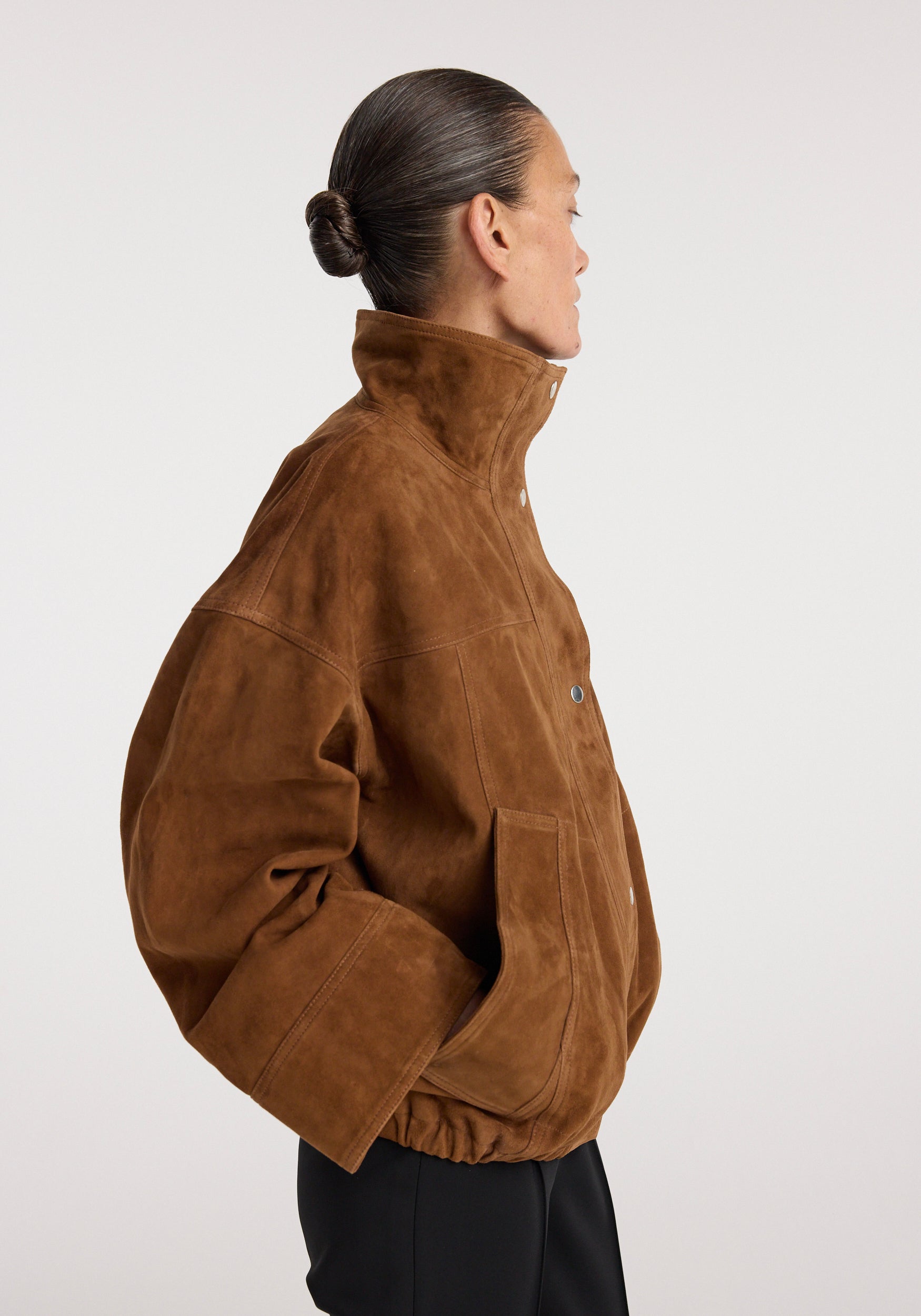 Woman wearing a cognac-colored Reinvented suede jacket | cognac with high collar and oversized sleeves, shown in profile against a neutral background.