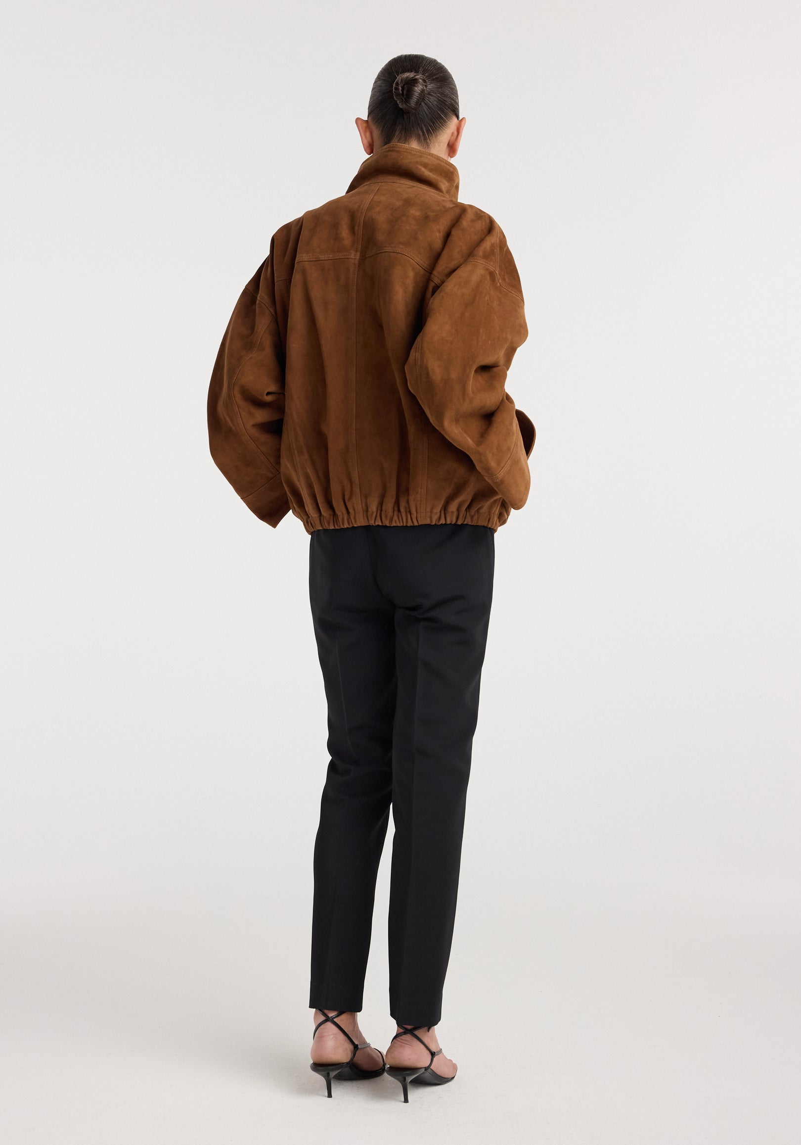 Woman seen from behind wearing a loose-fitting Reinvented suede jacket | cognac with a high collar, paired with black tailored pants and strappy heels.