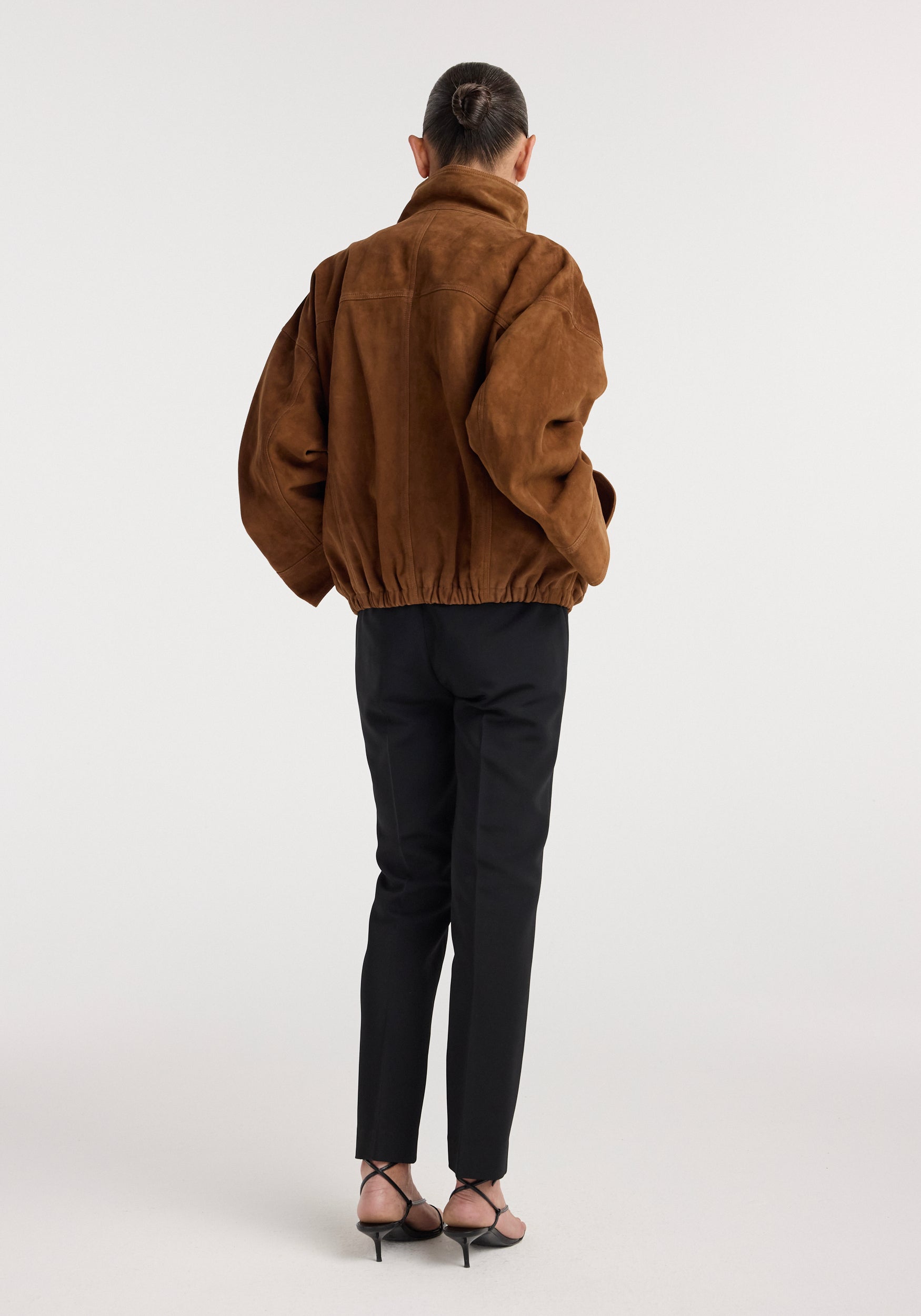 Woman seen from behind wearing a loose-fitting Reinvented suede jacket | cognac with a high collar, paired with black tailored pants and strappy heels.
