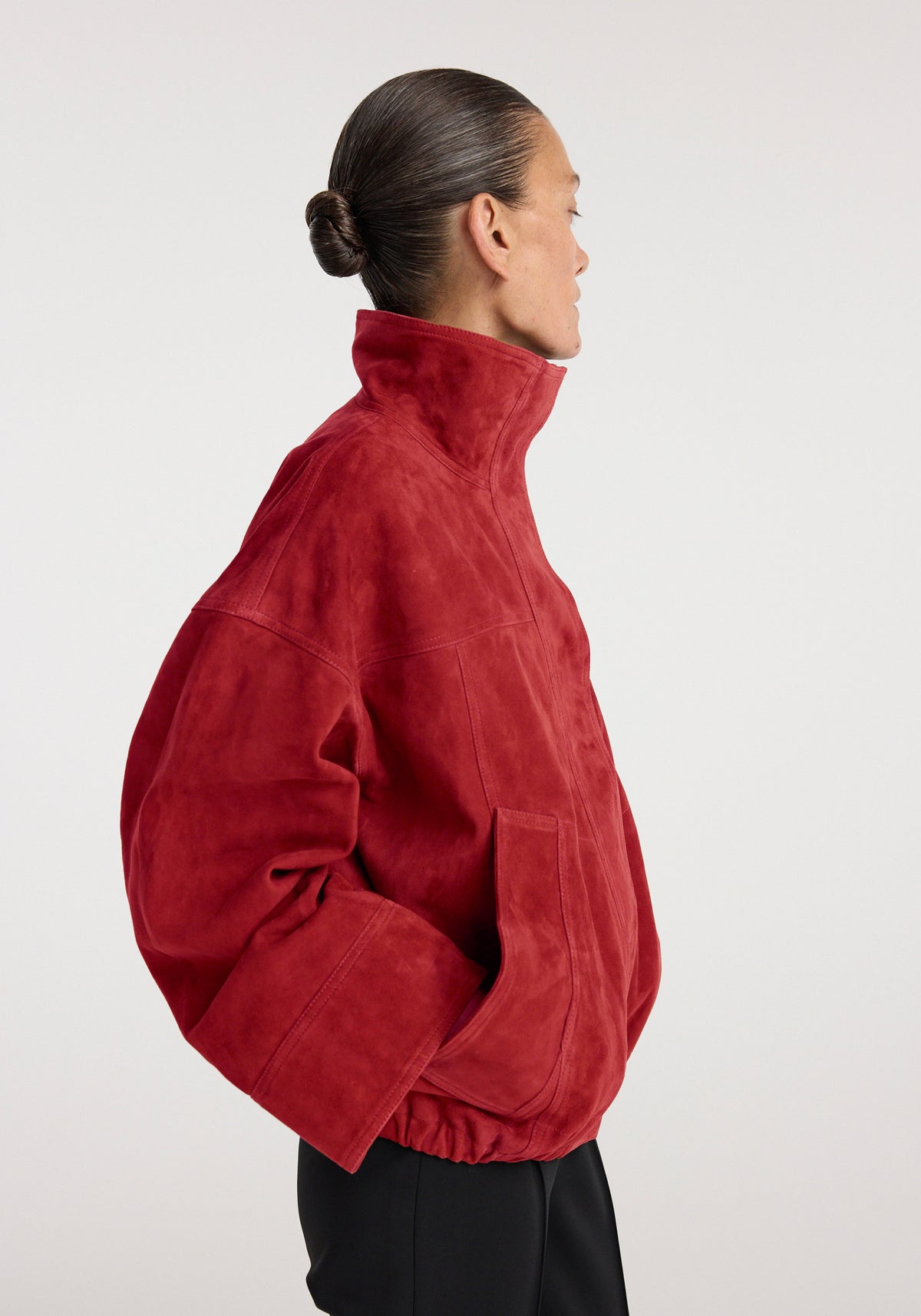 Side view of a person wearing a red Reinvented suede jacket | red with a high collar and loose fit, paired with black pants against a plain background.