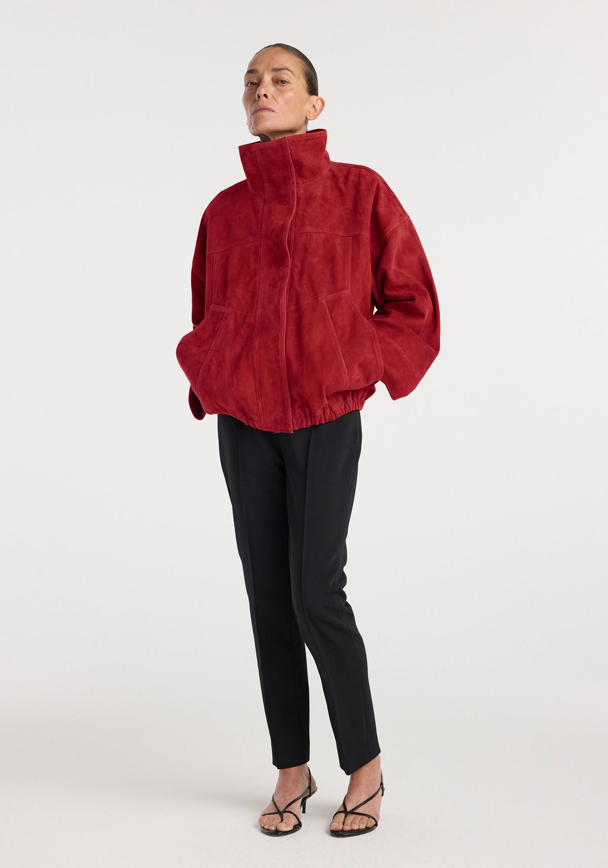 Woman wearing a high-collar, loose-fitting Reinvented suede jacket | red paired with black tapered pants and black strappy heels against a plain white background.
