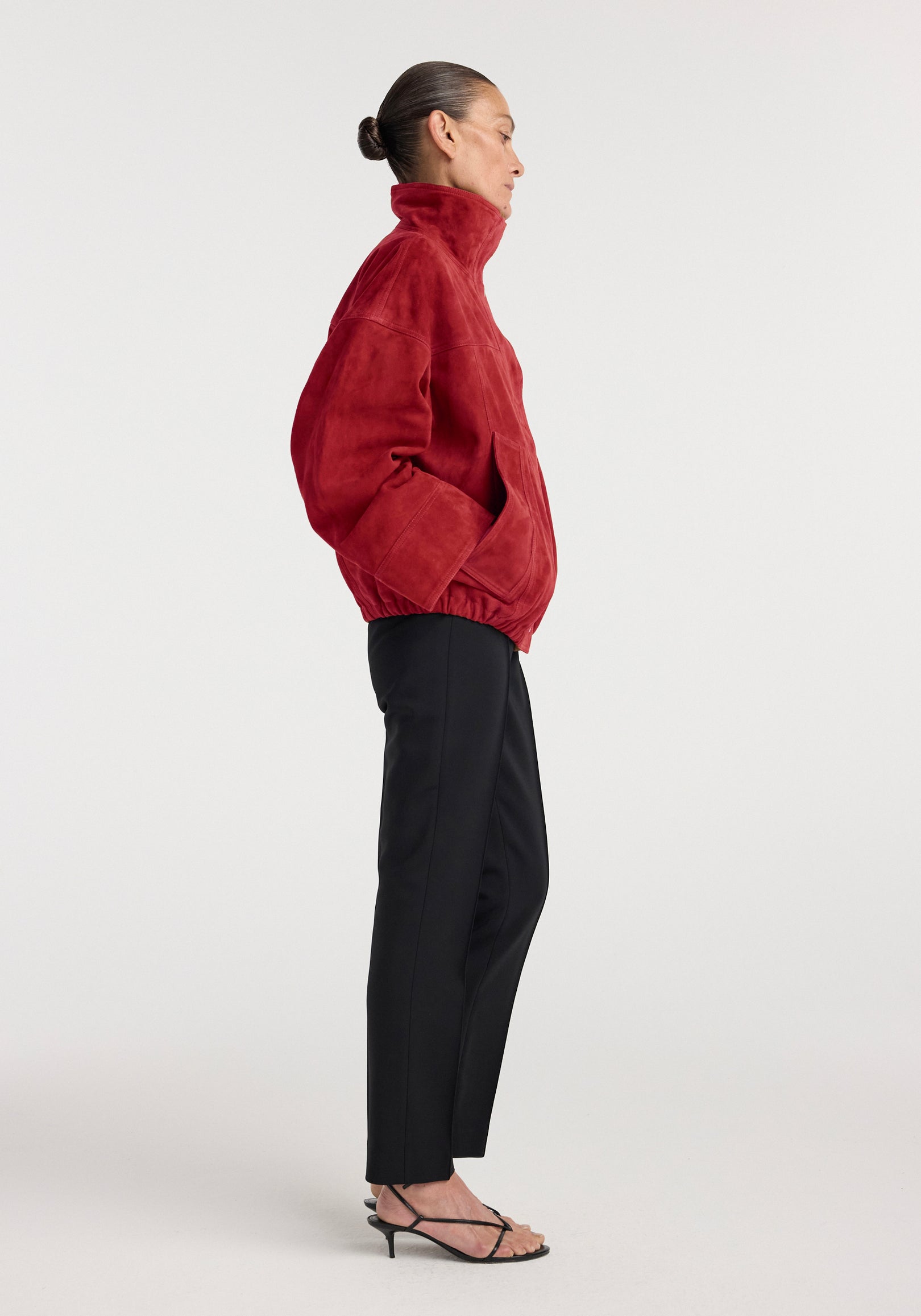 Side view of a woman wearing a red Reinvented suede jacket | red with a high collar, paired with black pants and strappy black heels against a plain background.