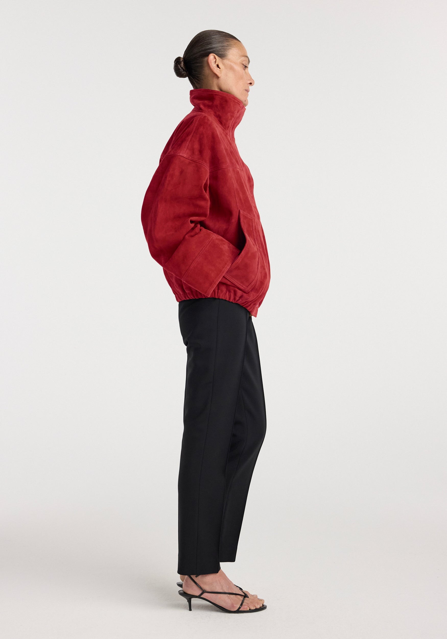 Side view of a woman wearing a red Reinvented suede jacket | red with a high collar, paired with black pants and strappy black heels against a plain background.