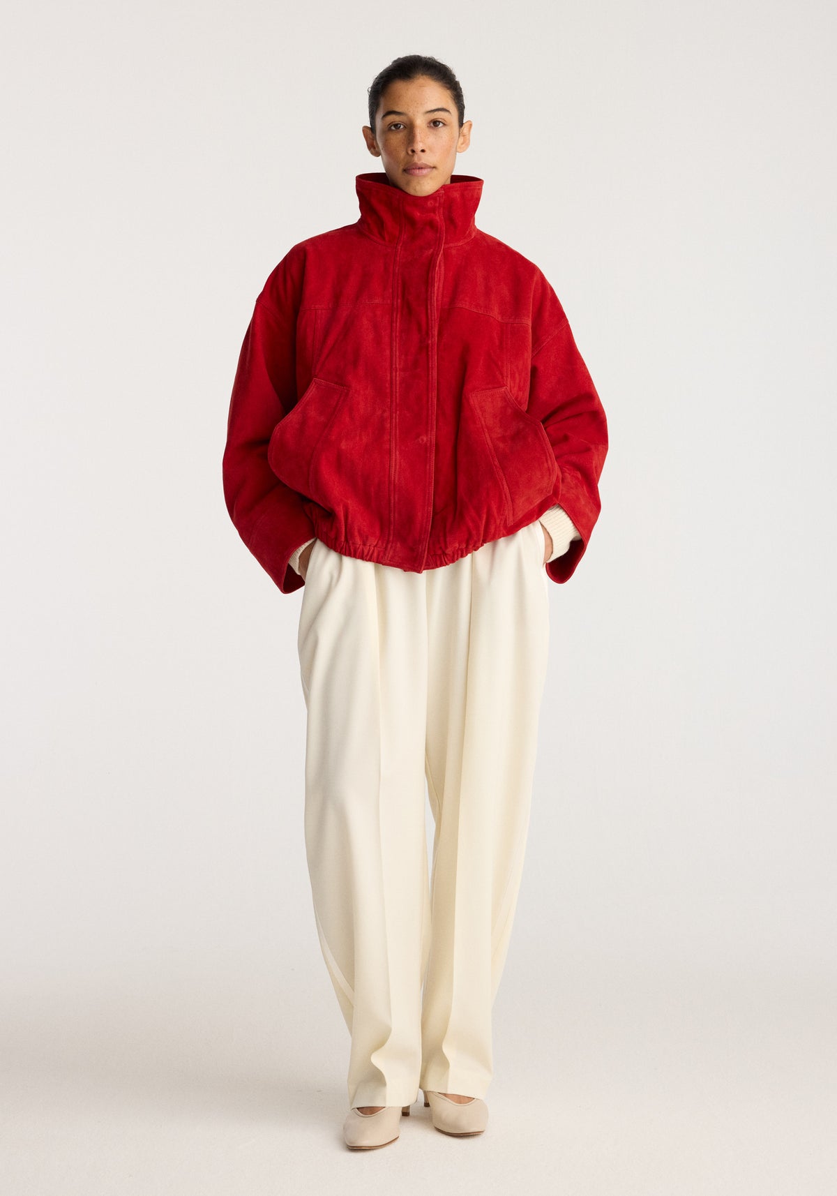Reinvented suede jacket | red worn by a model with a high stand collar, zip front, gathered hem, and spacious sleeves, styled with cream trousers.