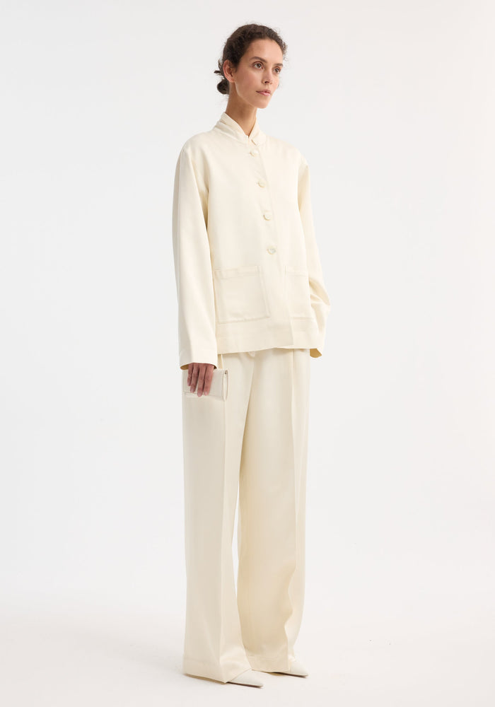Model wearing Mandarin pearl button satin jacket with matching wide-leg trousers, holding a neutral clutch, showcasing a minimalist, elegant ensemble against a plain background.