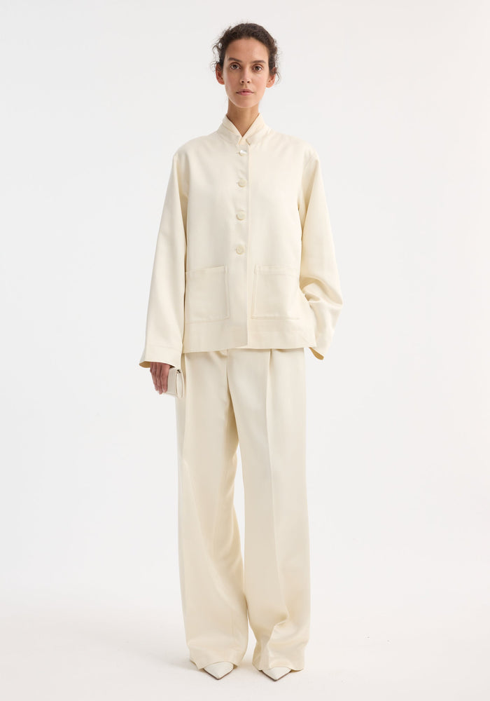Woman in Mandarin pearl button satin jacket, cream, with high-waisted trousers and clutch. Loose fit, mandarin collar, pearl buttons, front patch pockets.