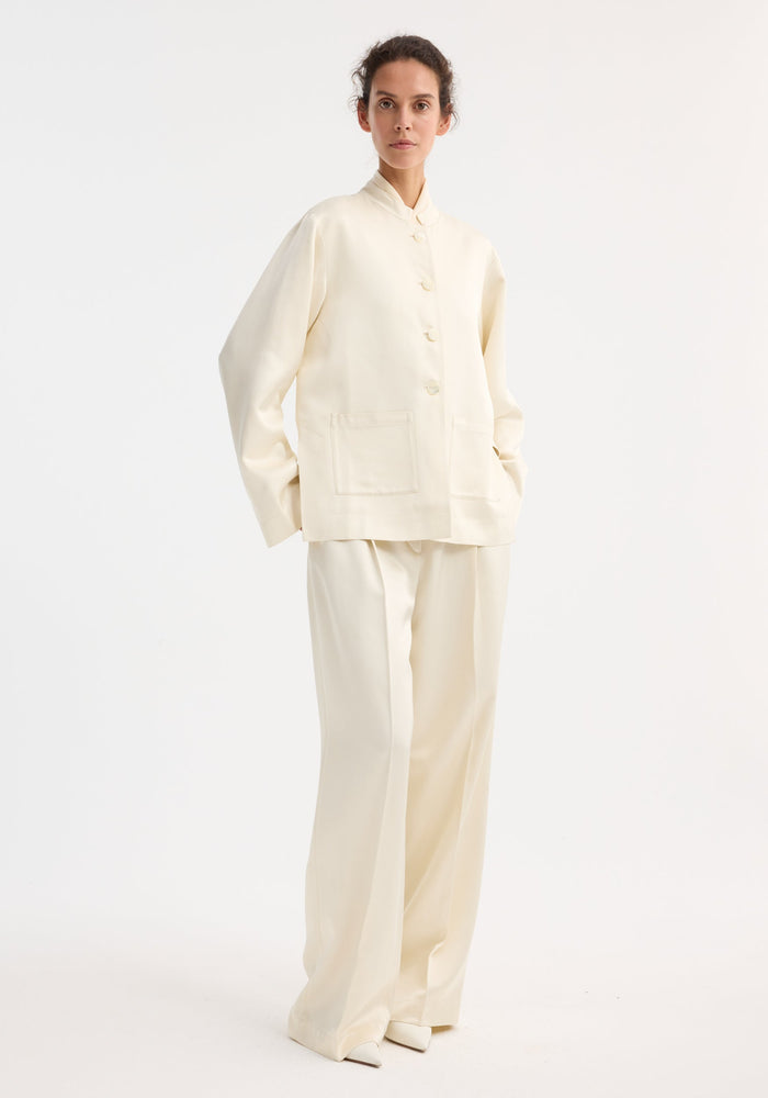 Model wearing Mandarin pearl button satin jacket with stand-up collar, pearl buttons, patch pockets, paired with matching wide-leg trousers, showcasing a refined, tonal ensemble.