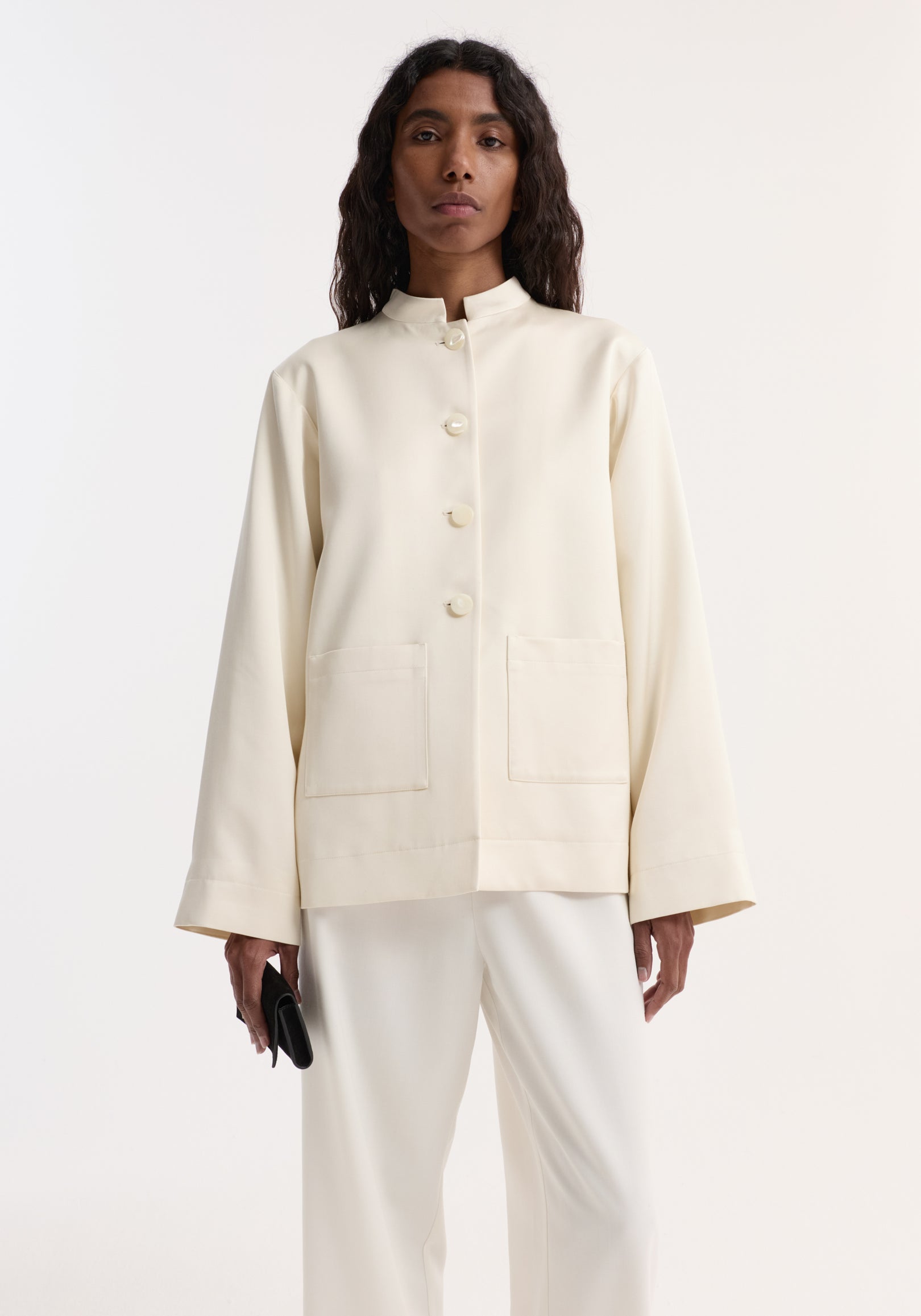 Woman wearing a cream satin jacket with pearl buttons and patch pockets, styled with matching cream pants and holding a small black clutch. Pearl button satin jacket | cream.