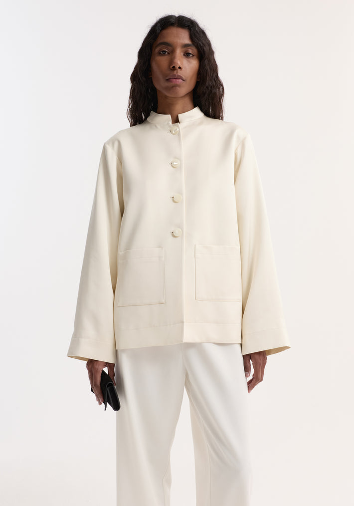 Model in Pearl button satin jacket | cream, featuring a mandarin collar, pearl buttons, and patch pockets, paired with white trousers and holding a black clutch.