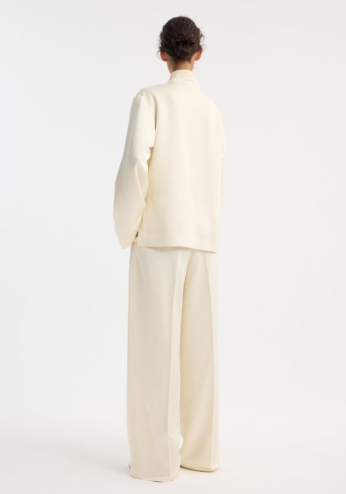 Person wearing Mandarin pearl button satin jacket with mandarin collar, front patch pockets, paired with matching wide-leg trousers. Hair in a simple bun.