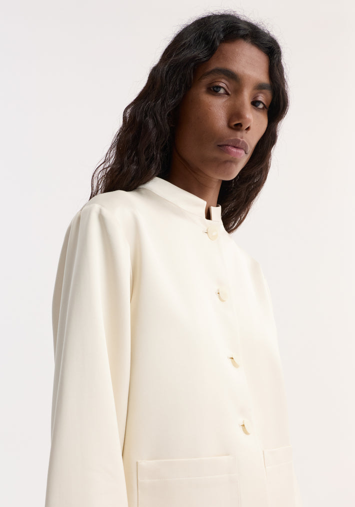 Pearl button satin jacket in cream with a stand-up collar, large patch pockets, and pearl buttons, modeled by a dark-haired person.
