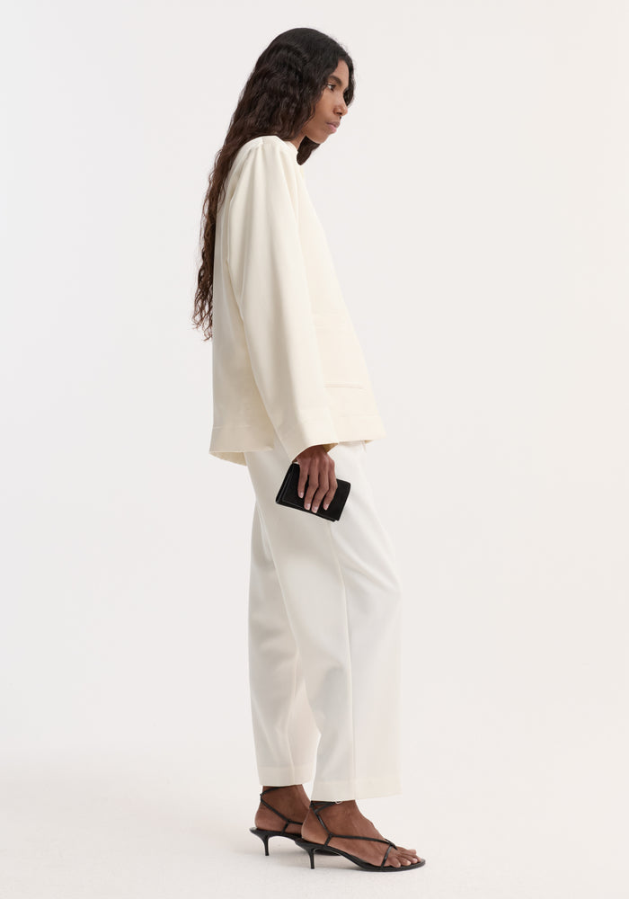 Model wearing a Pearl button satin jacket with stand-up collar, pearl buttons, and front patch pockets, paired with matching wide-leg trousers and black sandals, holding a wallet.