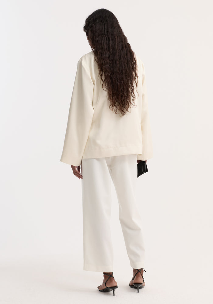 Back view of a person in a Pearl button satin jacket | cream, showcasing stand-up collar, pearl buttons, paired with matching wide-leg trousers, holding a black clutch.
