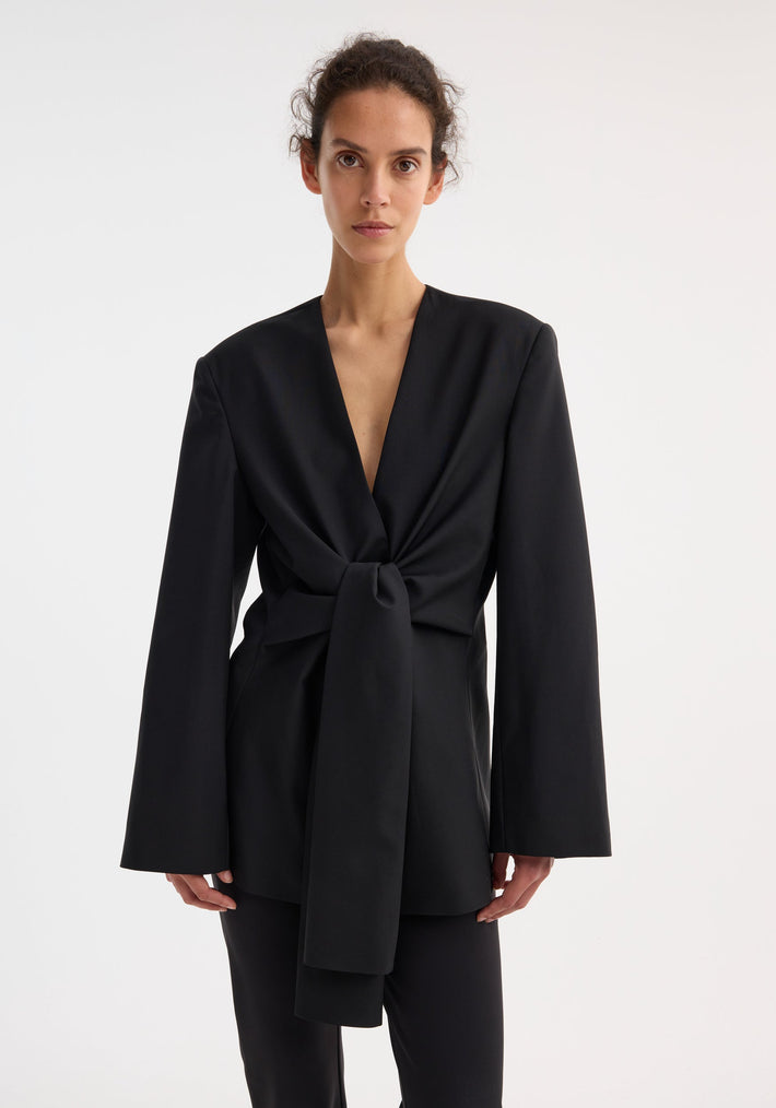 Sleeve-cinched blazer-top | black: Female model wearing a tailored wrap top with a deep V-neckline, flared sleeves, and a front knot detail, paired with slim trousers.