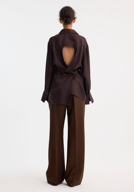 Model wearing Deconstructed silk shirt | mahogany, featuring an open back with tie detail, matched with wide-leg trousers and beige pointed-toe shoes.