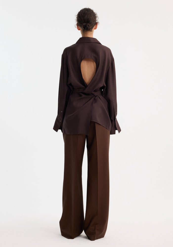 Model wearing Deconstructed silk shirt in mahogany, showcasing open back with tie detail, paired with matching wide-leg trousers, highlighting the shirt's elegant, versatile style.
