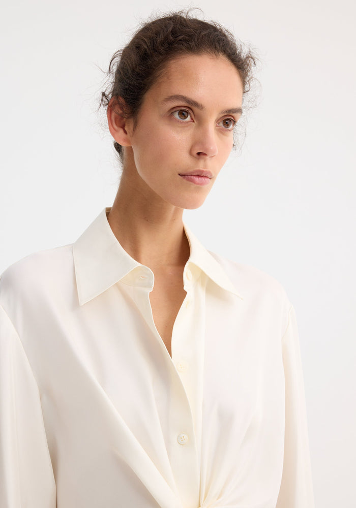 Young woman wearing a Silk knotted shirt | cream, showcasing a twisted tie front design and classic pointed collar, gazing slightly off to one side.