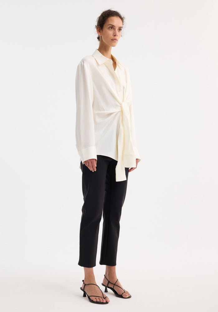 Silk knotted shirt | cream features a long-sleeve blouse with a twisted tie front, paired with tailored black trousers and open-toe sandals on a model.