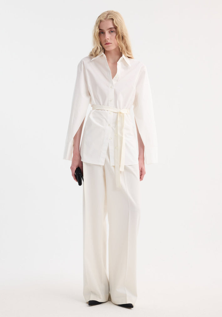 Model in Poplin silk combined gilet-shirt with open side-slit sleeves, wide-leg trousers, black clutch, and shoes; features cotton front, silk back.