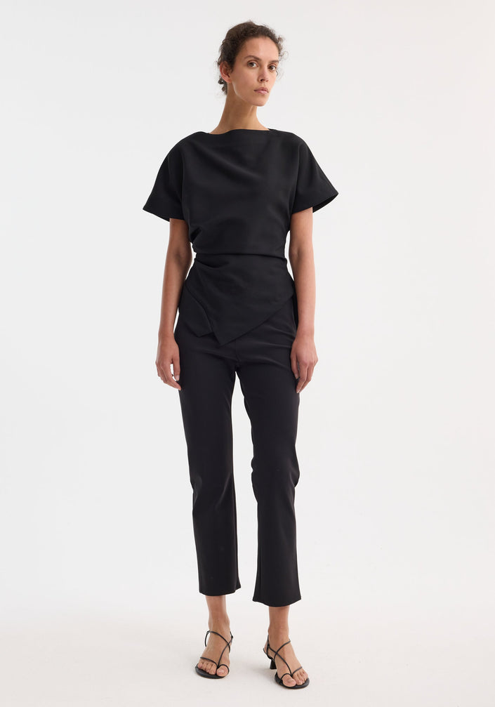 Model wearing Draped lapel top with sculptural asymmetrical silhouette, featuring a slashed neckline and deconstructed lapel, paired with slim-fit pants and strappy sandals.