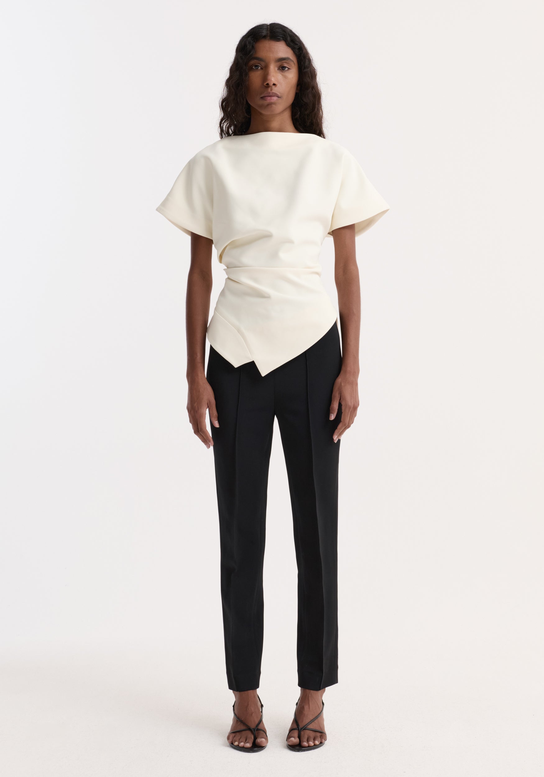Woman wearing a draped lapel top | off white with short sleeves and an asymmetrical waist detail, paired with black tailored pants and strappy black heels.