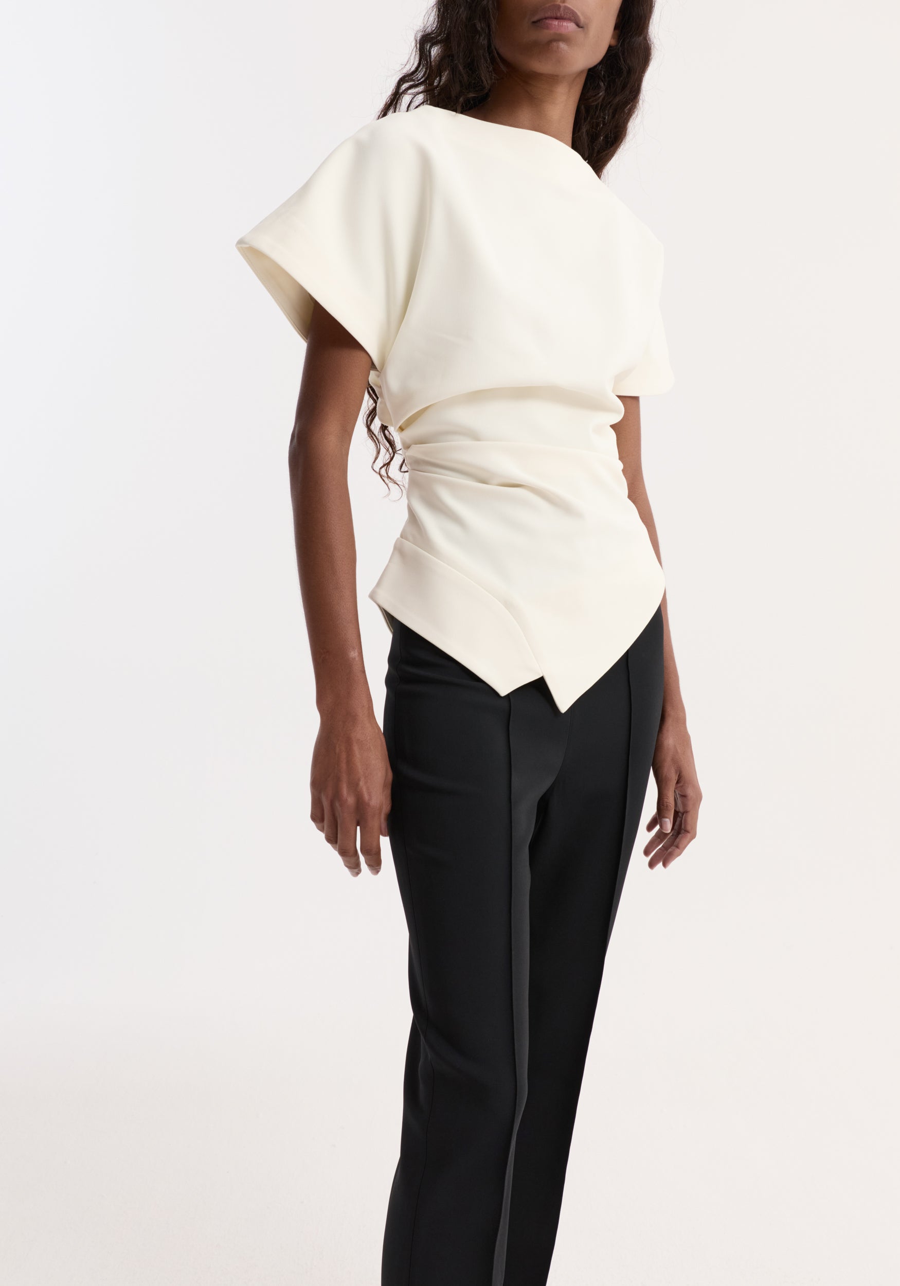 Woman wearing a draped lapel top | off white with short wide sleeves and structured waist detail, paired with black tailored pants against a plain background.