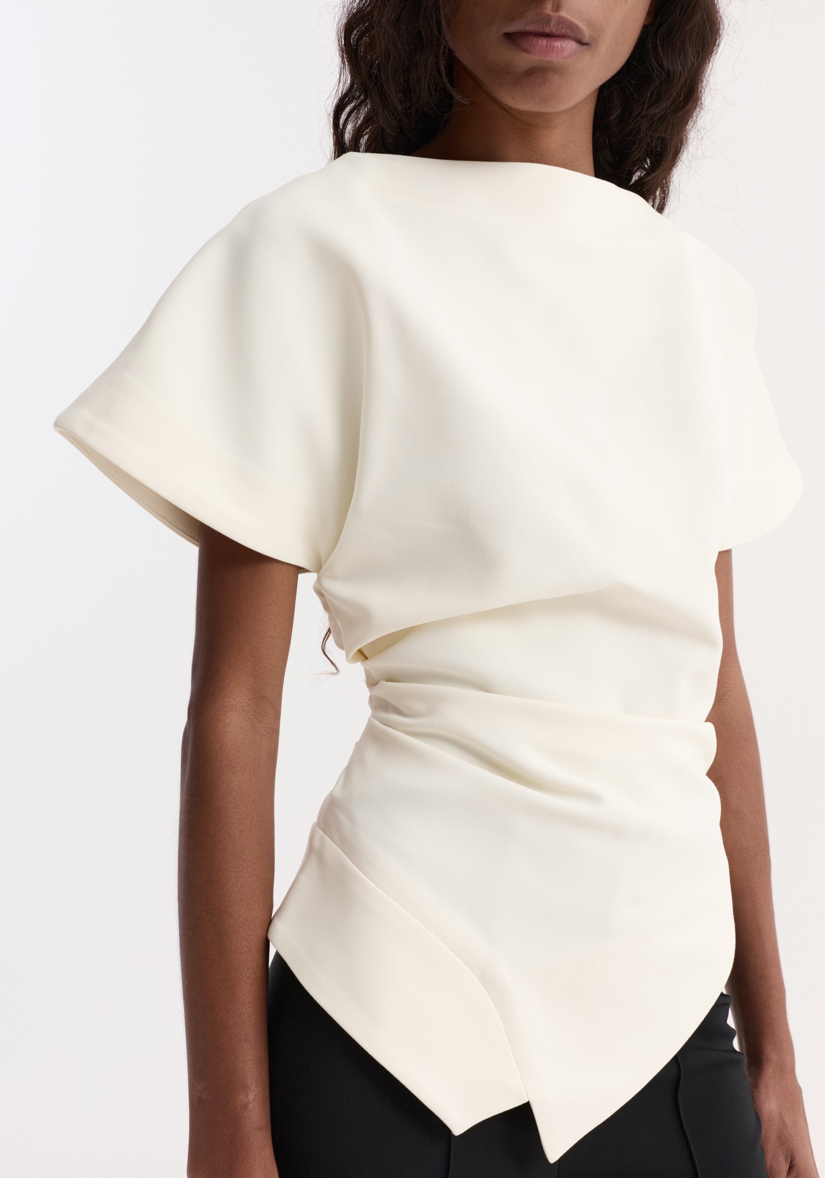 Close-up of a woman wearing a draped lapel top | off white, featuring short sleeves, a boat neckline, and a tailored, asymmetrical hem.