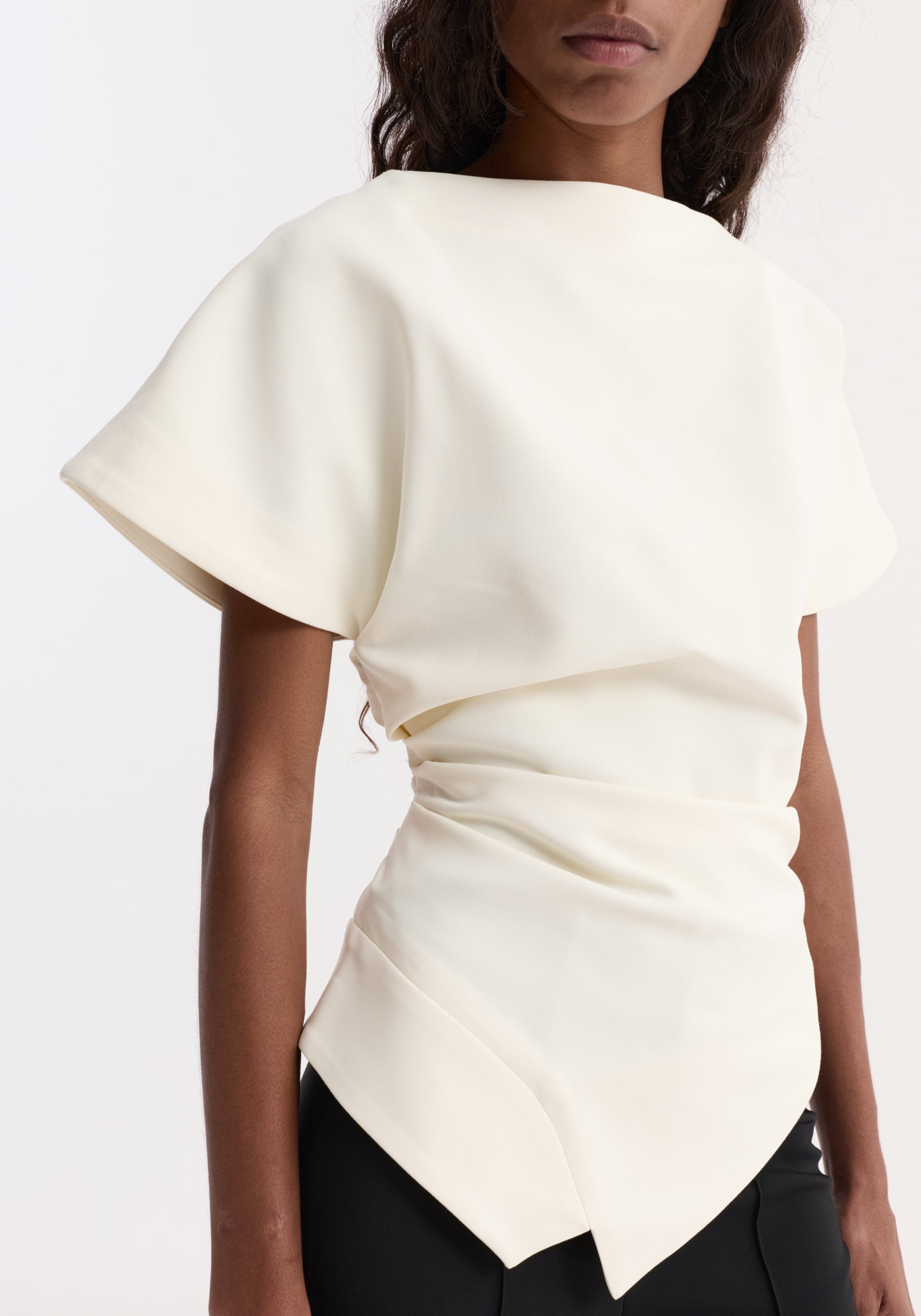 Close-up of a woman wearing a draped lapel top | off white, featuring short sleeves, a boat neckline, and a tailored, asymmetrical hem.