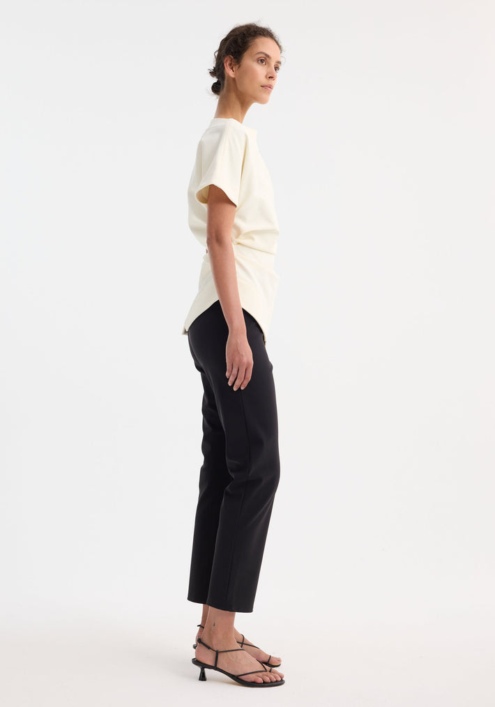 Woman in profile wearing Draped lapel top in off white, featuring a slashed neckline and sculpted shoulder, paired with black trousers and strappy sandals.