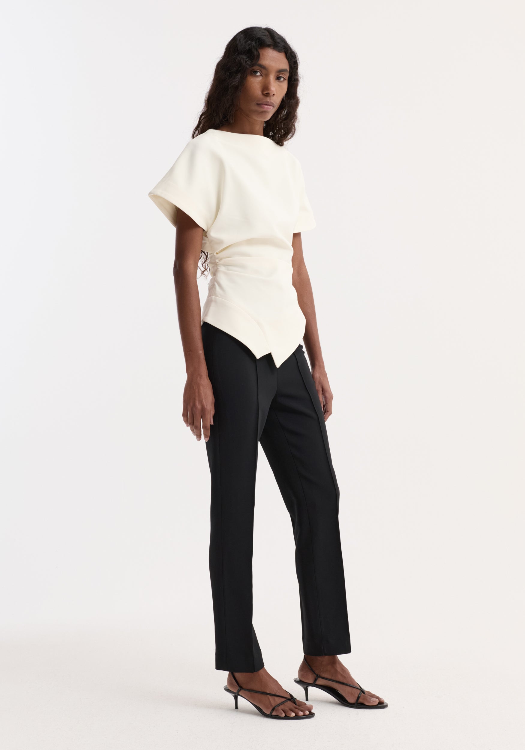 Woman wearing a draped lapel top | off white with short sleeves, paired with black slim trousers and strappy low-heeled sandals against a plain background.