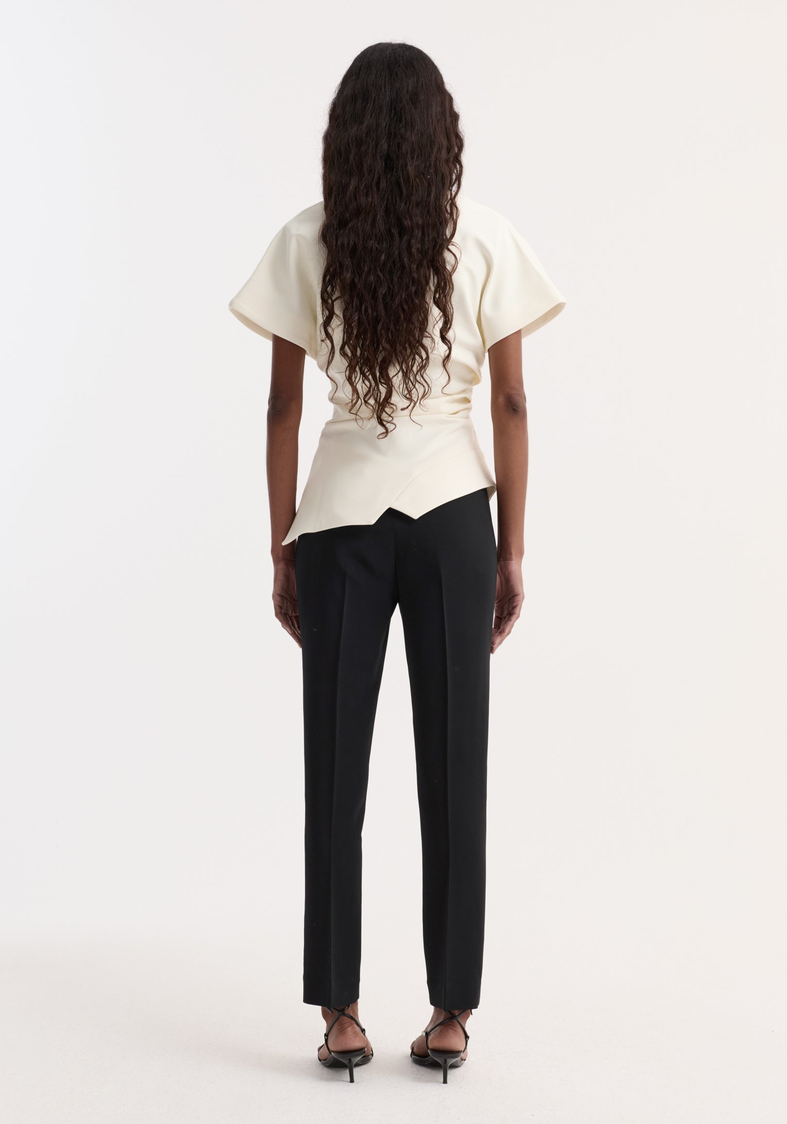 Woman wearing off white draped lapel top with short sleeves paired with black tailored trousers and black strappy heels, shown from the back.