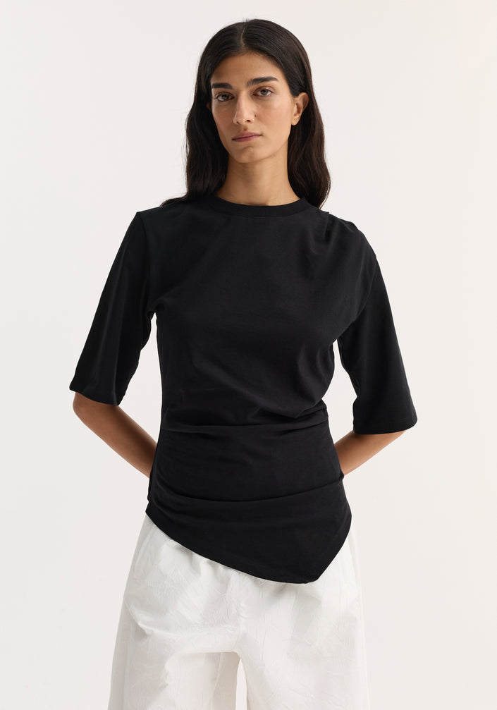 Woman wearing Reinvented twisted t-shirt | black, showcasing its fitted, asymmetrical twist design, paired with loose white trousers and black heeled sandals.