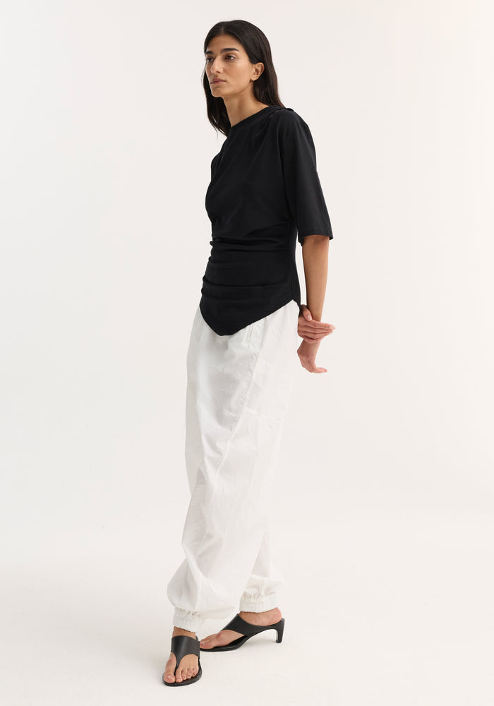 Model wears the Reinvented twisted t-shirt with an asymmetrical, ruched hem, paired with white ankle-length jogger-style pants and black open-toe mules.