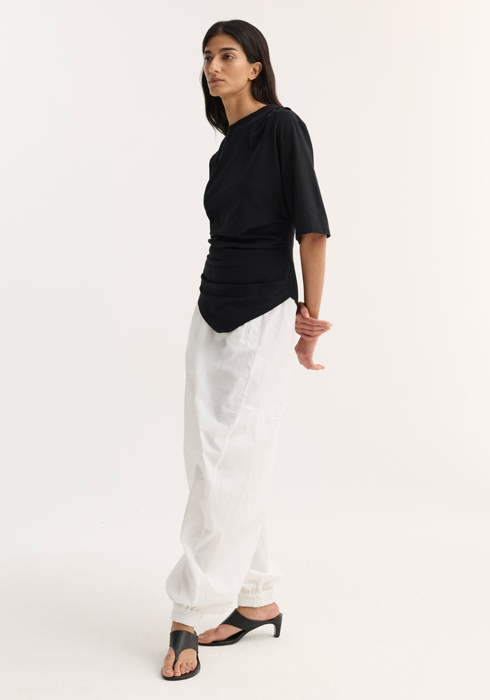 Model wears the Reinvented twisted t-shirt with an asymmetrical, ruched hem, paired with white ankle-length jogger-style pants and black open-toe mules.