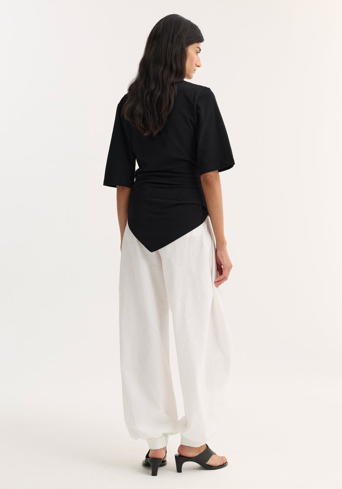 Reinvented twisted t-shirt in black, featuring a sculptural, asymmetrical drape with elbow-length sleeves, paired with white ankle-gathered trousers and black mule sandals.
