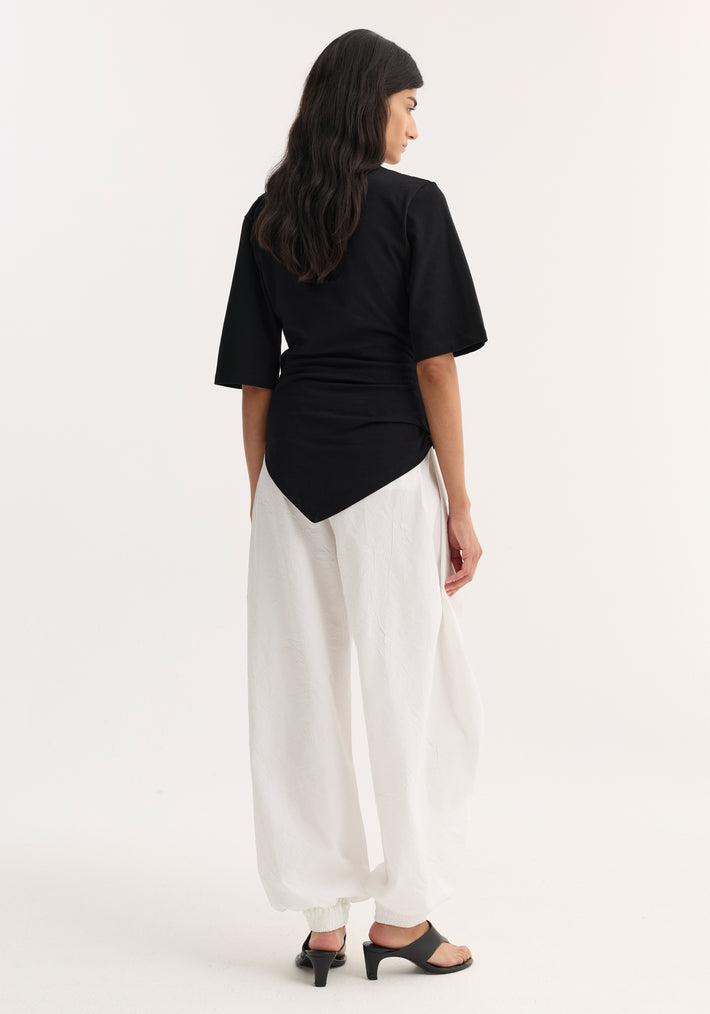 Reinvented twisted t-shirt in black, featuring a sculptural, asymmetrical drape with elbow-length sleeves, paired with white ankle-gathered trousers and black mule sandals.